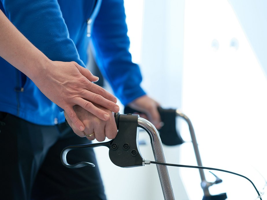 Ein Patient geht mit einem Rollator und eine Pflegeassistentin hält seine Hand.